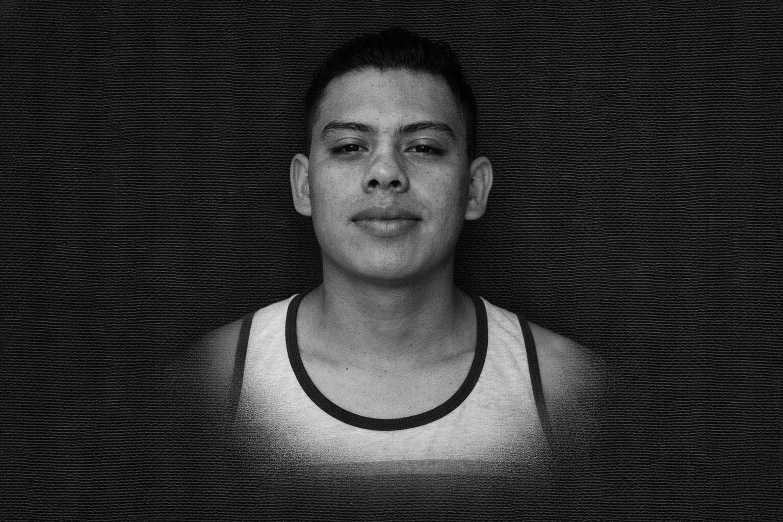 Retrato en blanco y negro de un hombre joven con camiseta sin mangas sobre un fondo negro texturizado.