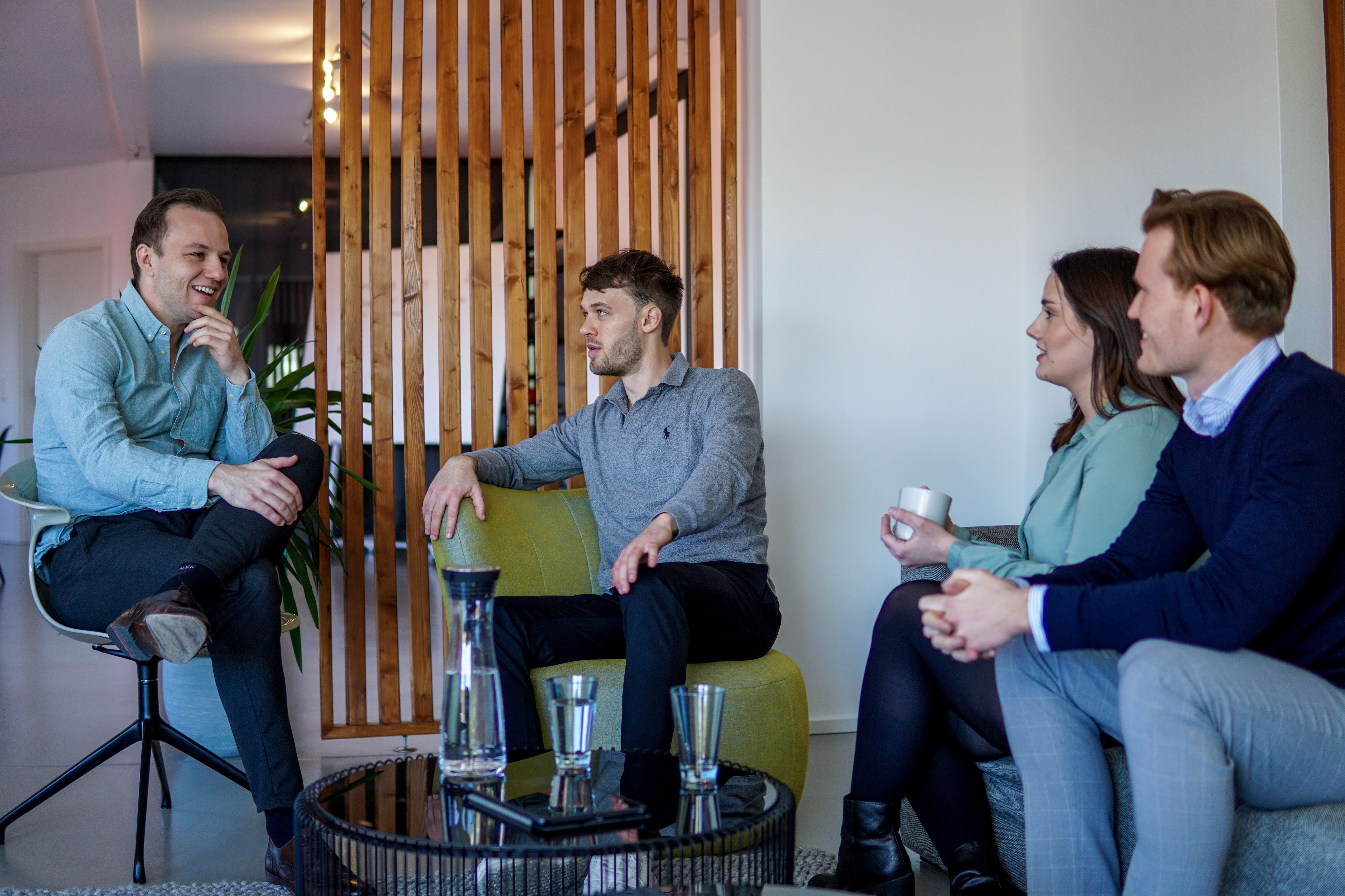 Eine Gruppe von vier jungen Erwachsenen sitzt in einer modernen Lounge- oder Büroumgebung und unterhält sich entspannt. Sie sitzen auf Hockern und Sesseln um einen niedrigen Tisch und trinken Kaffee oder Tee.