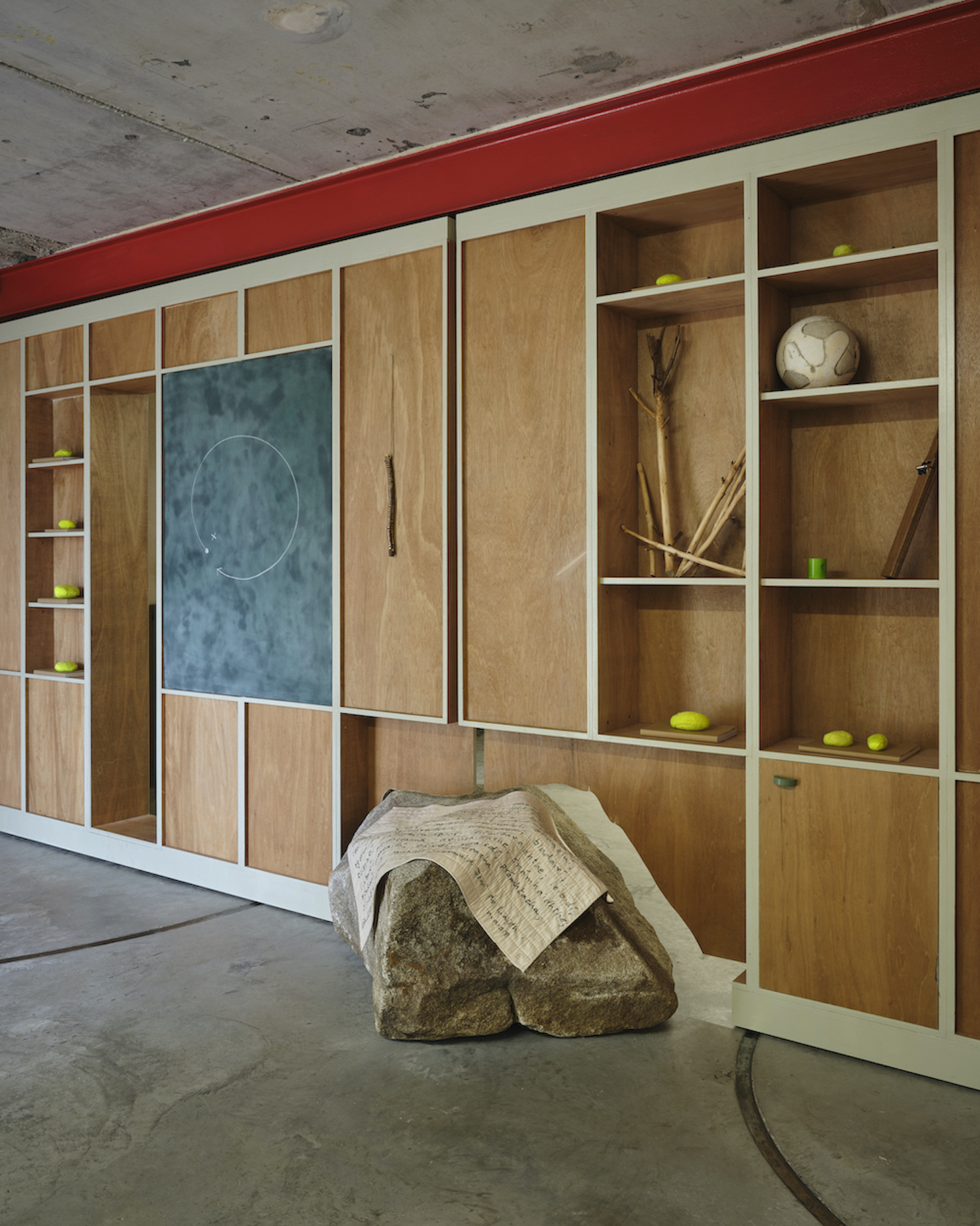 A wall of plywood shelving hovers over a granite boulder, placed on the concrete floor with a cloth draped over it. 