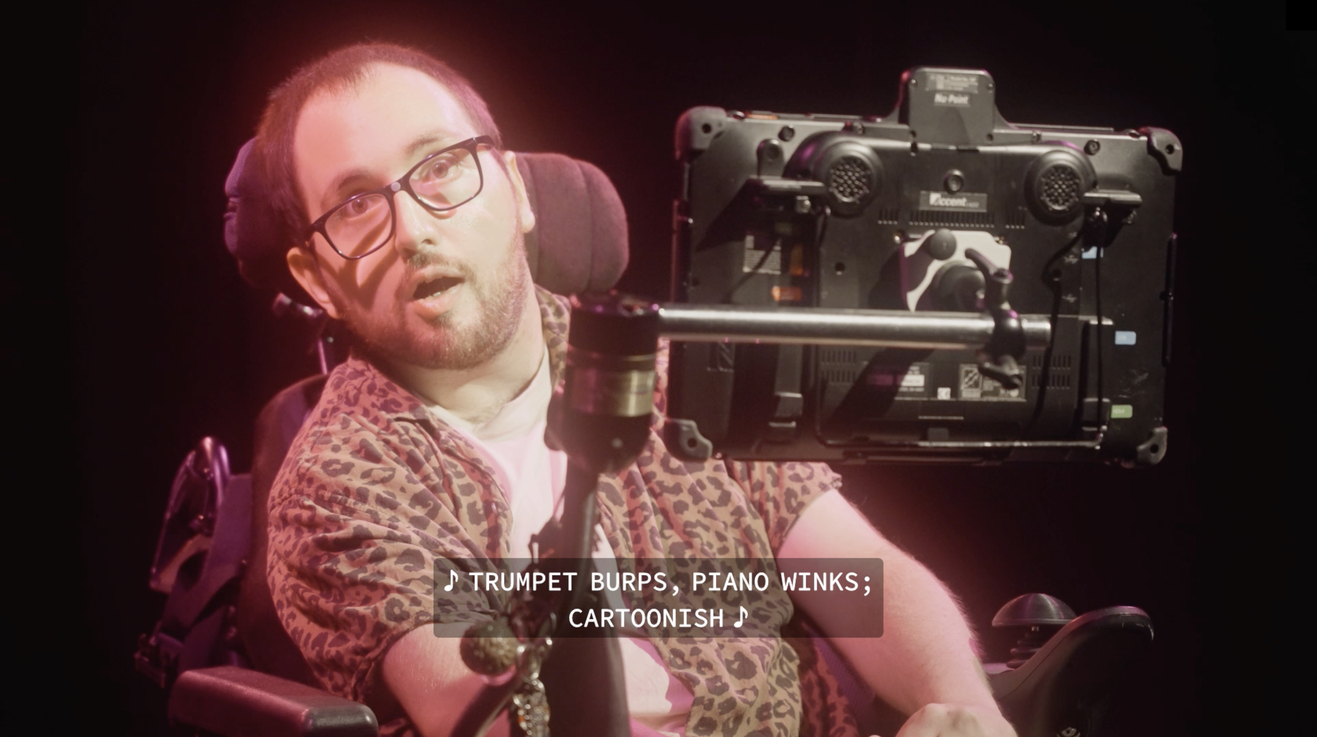 A young male performer seated in a wheelchair and behind a screen wears glasses and it lit in pink stage lights.