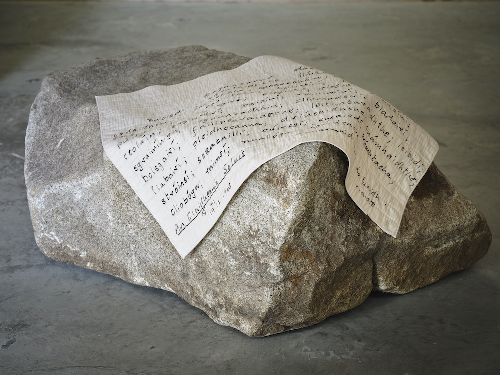 A large granite boulder sits on a concrete floor, with a piece of quilted cloth draped over it. Enlarged lines of handwriting are visible on the quilt.