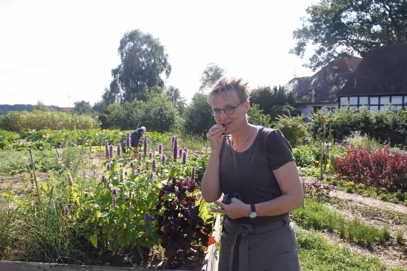 Person wearing glasses eating a plant in a sunny garden with another person working in the background.