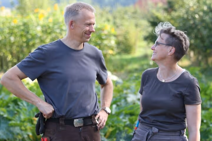 Middle-aged man and woman smiling and looking at each other outdoors with green plants in background.