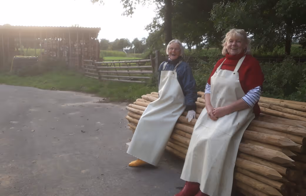 Two women wearing aprons and boots sitting on stacked wooden logs outdoors near a shed and trees.