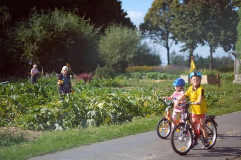 Two children wearing helmets riding bicycles on a path near a green garden with adults in the background.