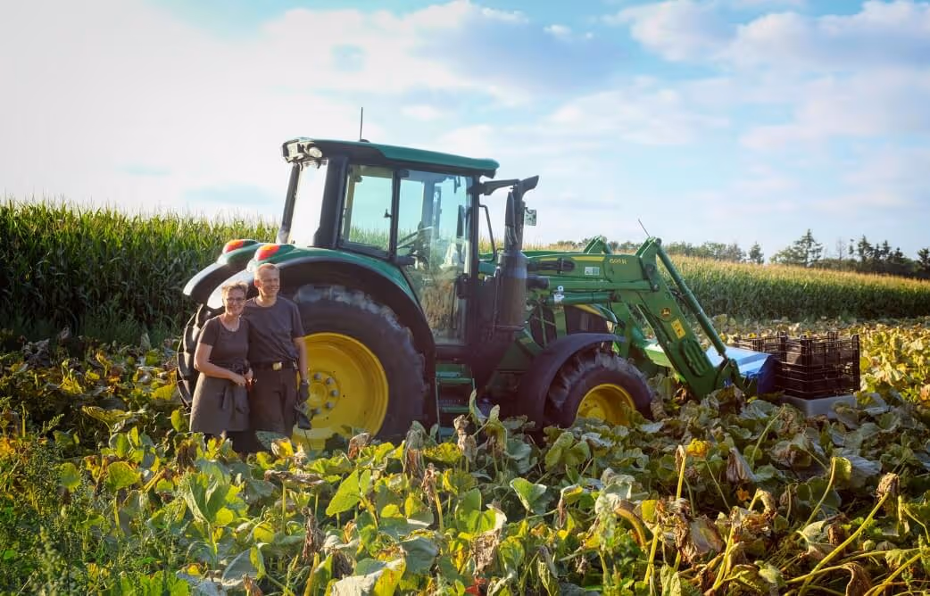 Two farmers standing beside a green tractor in a field with crops and a cornfield in the background.