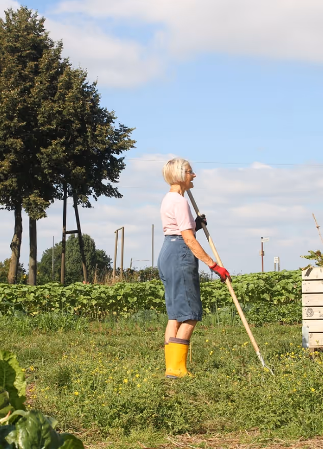Older woman wearing yellow boots gardening in a green field under blue sky with scattered clouds.