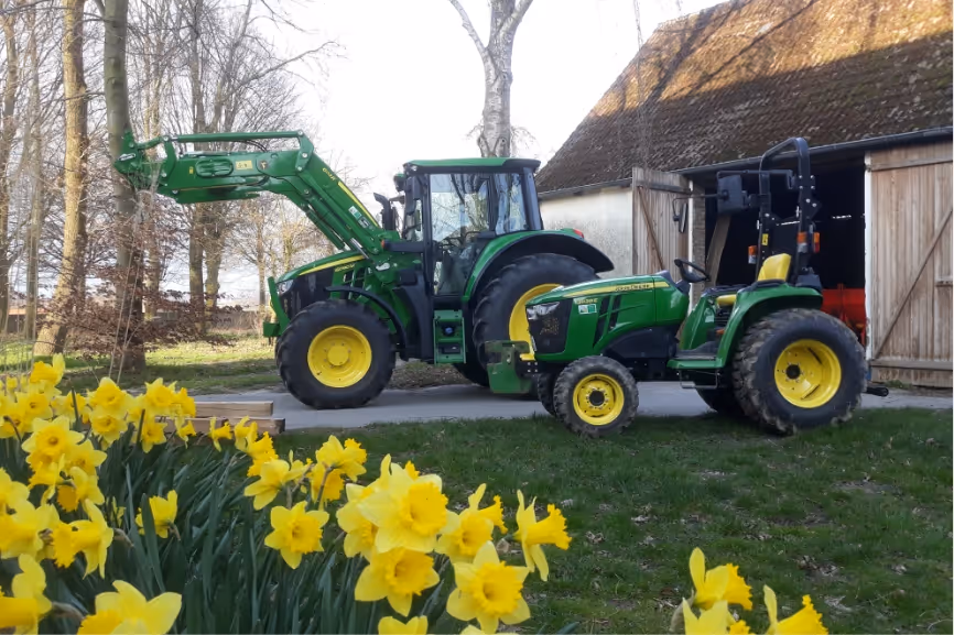 Two green John Deere tractors parked near a barn with yellow daffodils blooming in front.