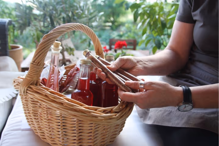 Person holding two glass tubes with spices over a basket of bottled red liquids and dried chili peppers.