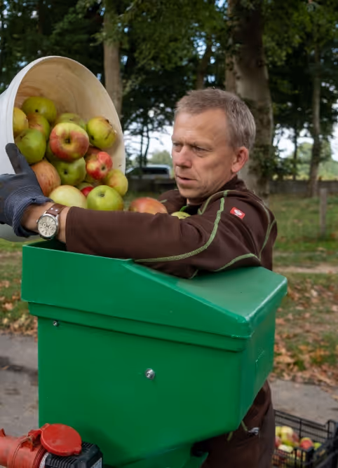 Man wearing gloves pouring apples from a wooden basket into a green apple grinder outdoors.