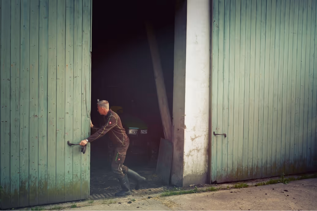 Man in work clothes pushing open large green wooden barn doors with tractor inside dimly lit barn.