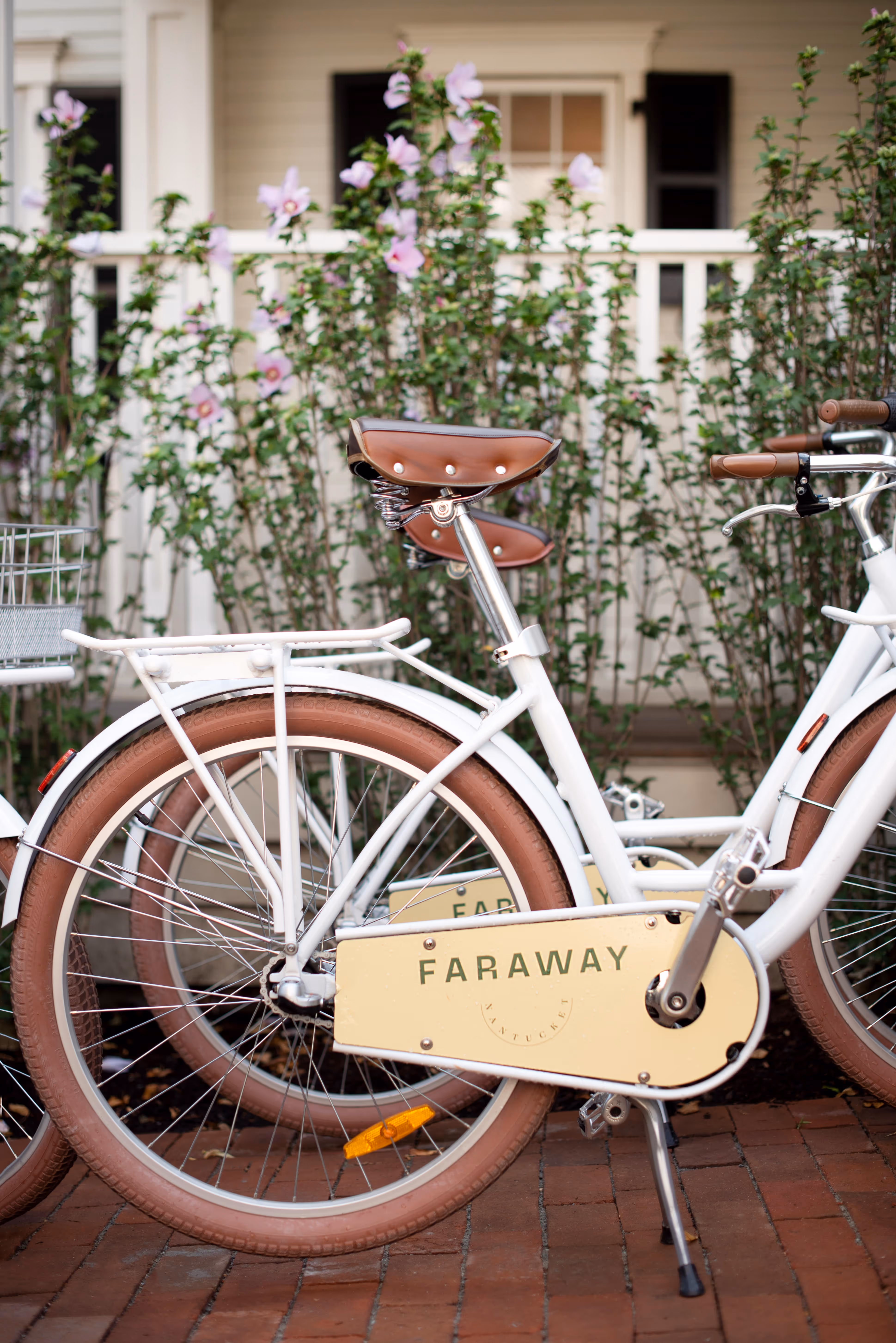 A white bicycle with tan tires and a FARAWAY sign is parked on a brick path in front of a white house with flowers.