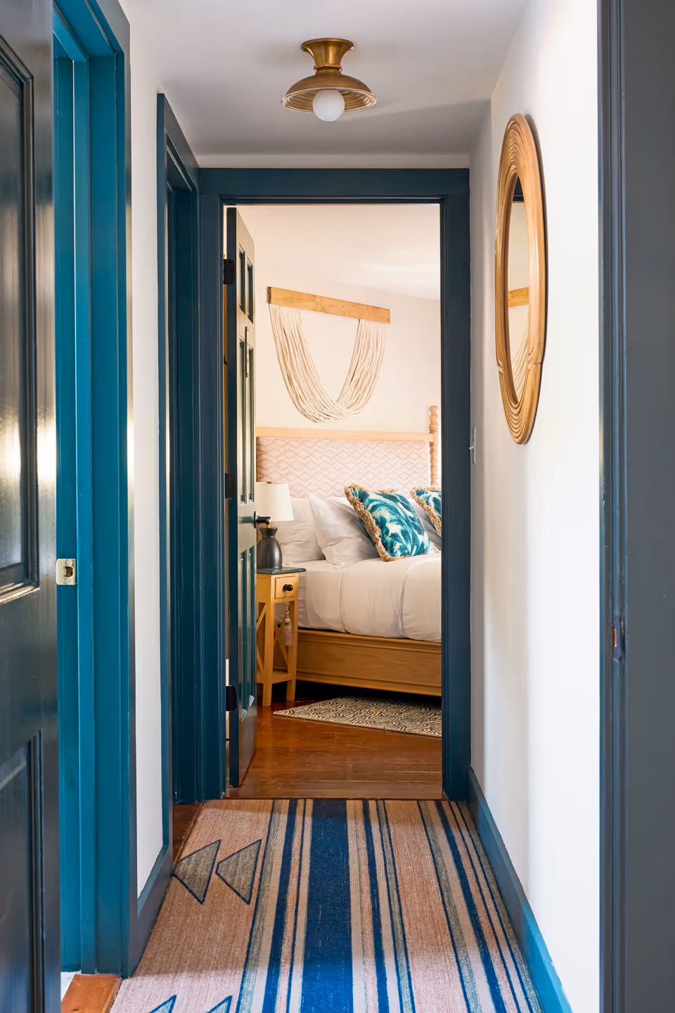A hallway with teal door frames and a striped rug leading into a cozy bedroom. The bedroom features a wooden bed with a patterned headboard, white bedding, and teal accent pillows. A macrame wall hanging decorates the wall above the bed, and a round wooden mirror hangs on the hallway wall.