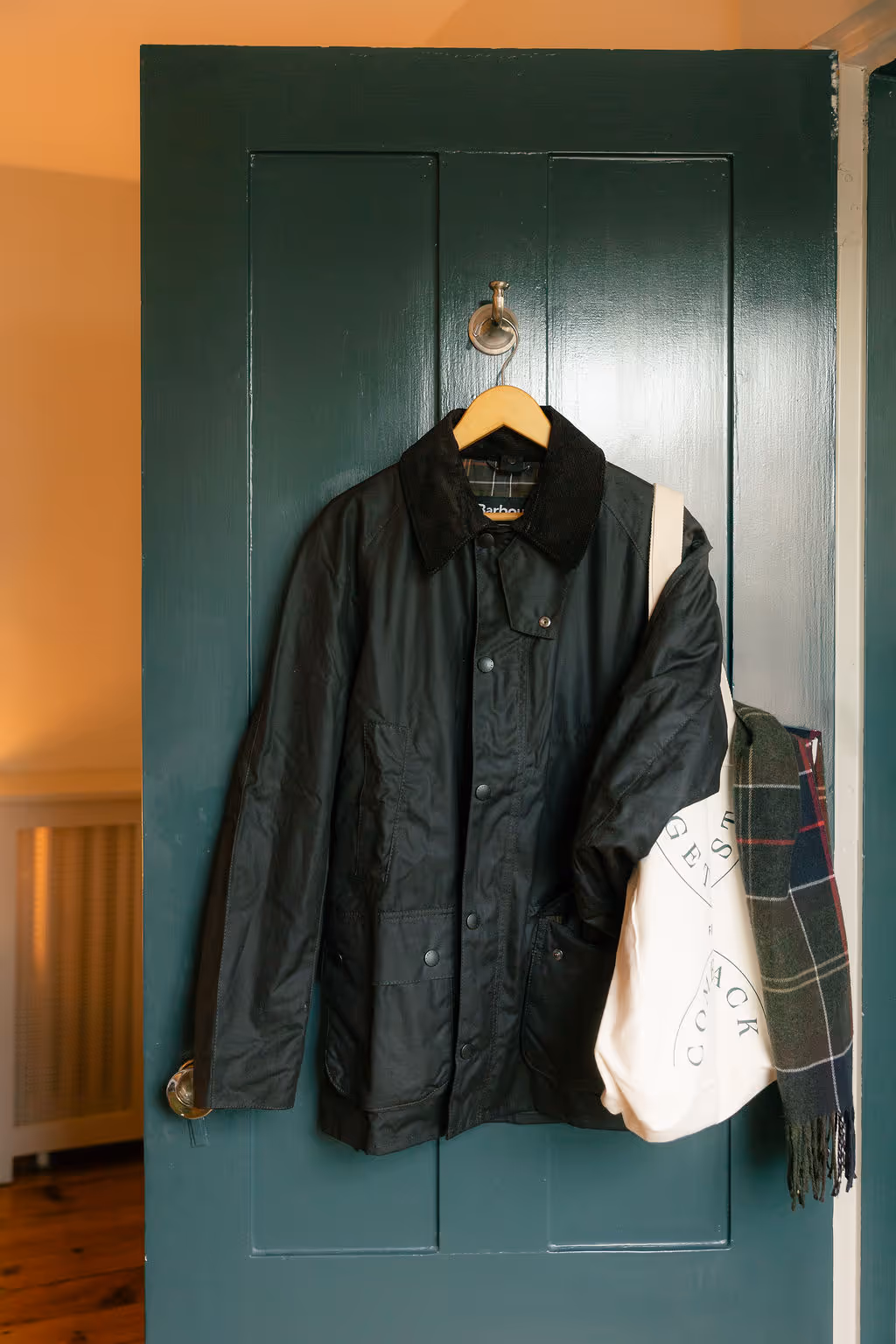 A black jacket, tote bag, and plaid scarf hang on a hook attached to a green door.