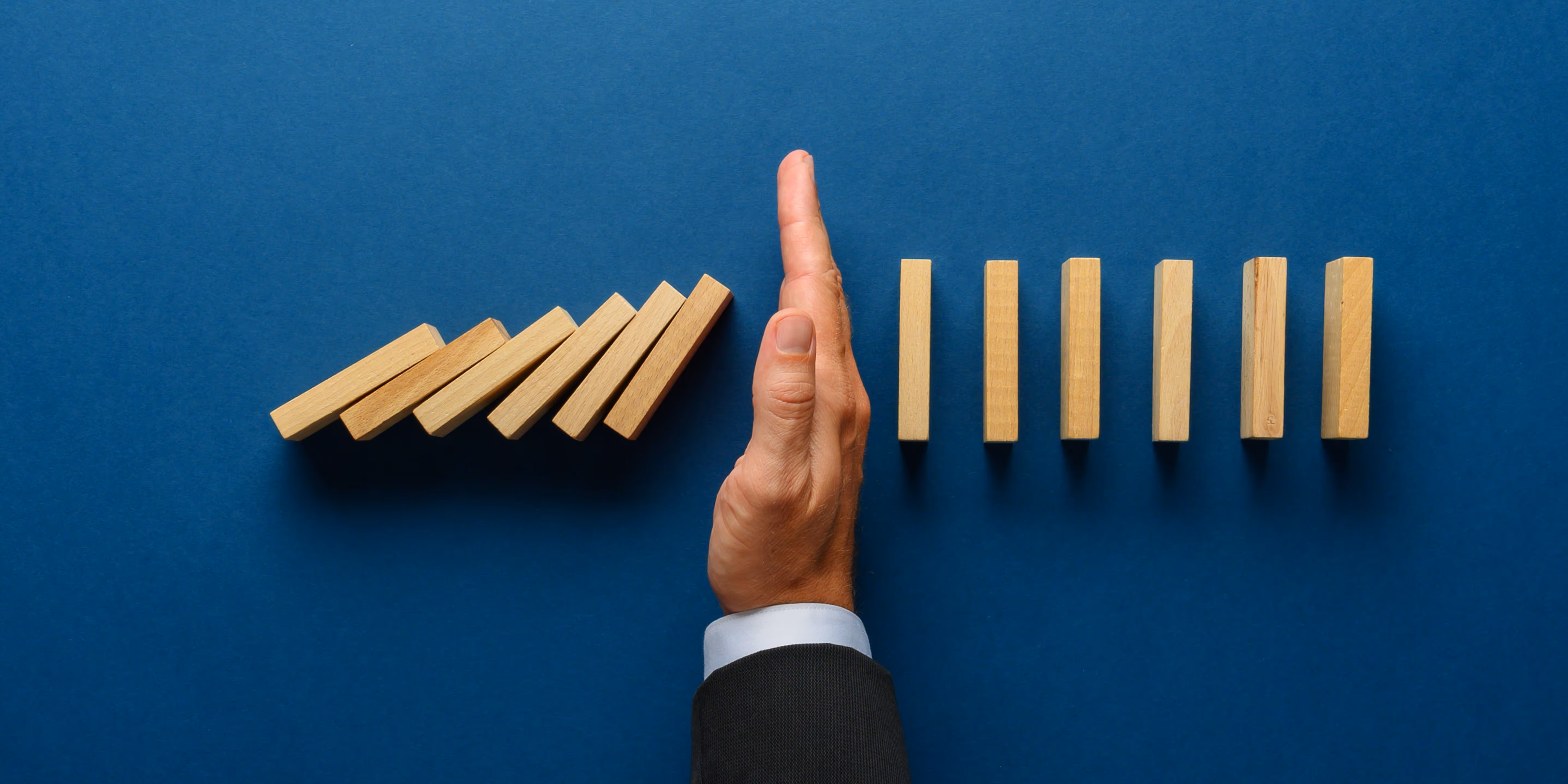 A set of wooden blocks that are falling over with an outstretched hand stopping them and a row of upright wooden blocks beyond the hand