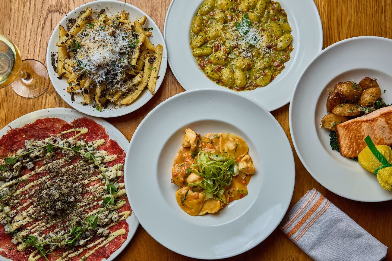Overhead view of an elegant Italian-inspired dinner spread on a wooden table, featuring truffle fries topped with parmesan, gnocchi in a creamy pesto sauce, beef carpaccio with microgreens, ravioli in tomato cream sauce, and a grilled salmon fillet with roasted potatoes and spinach, accompanied by a glass of white wine.
