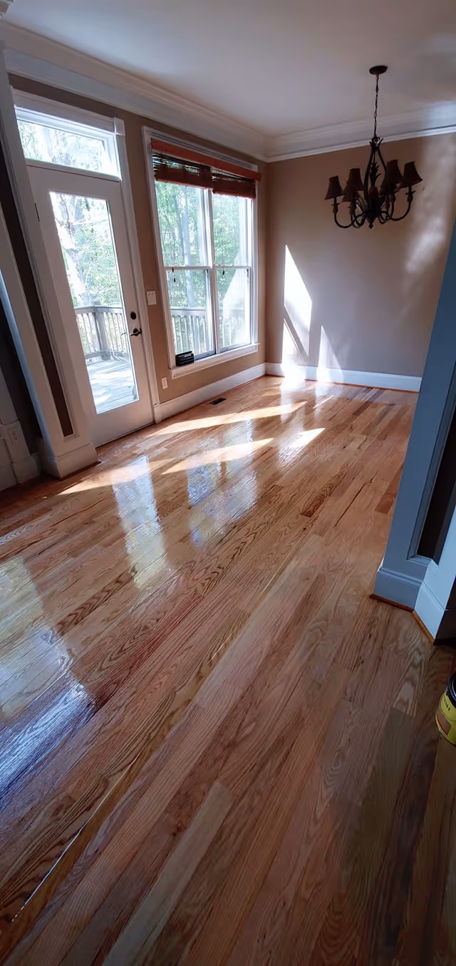 Sunlit empty room with polished hardwood floors, beige walls, large window, glass door, and a dark chandelier.
