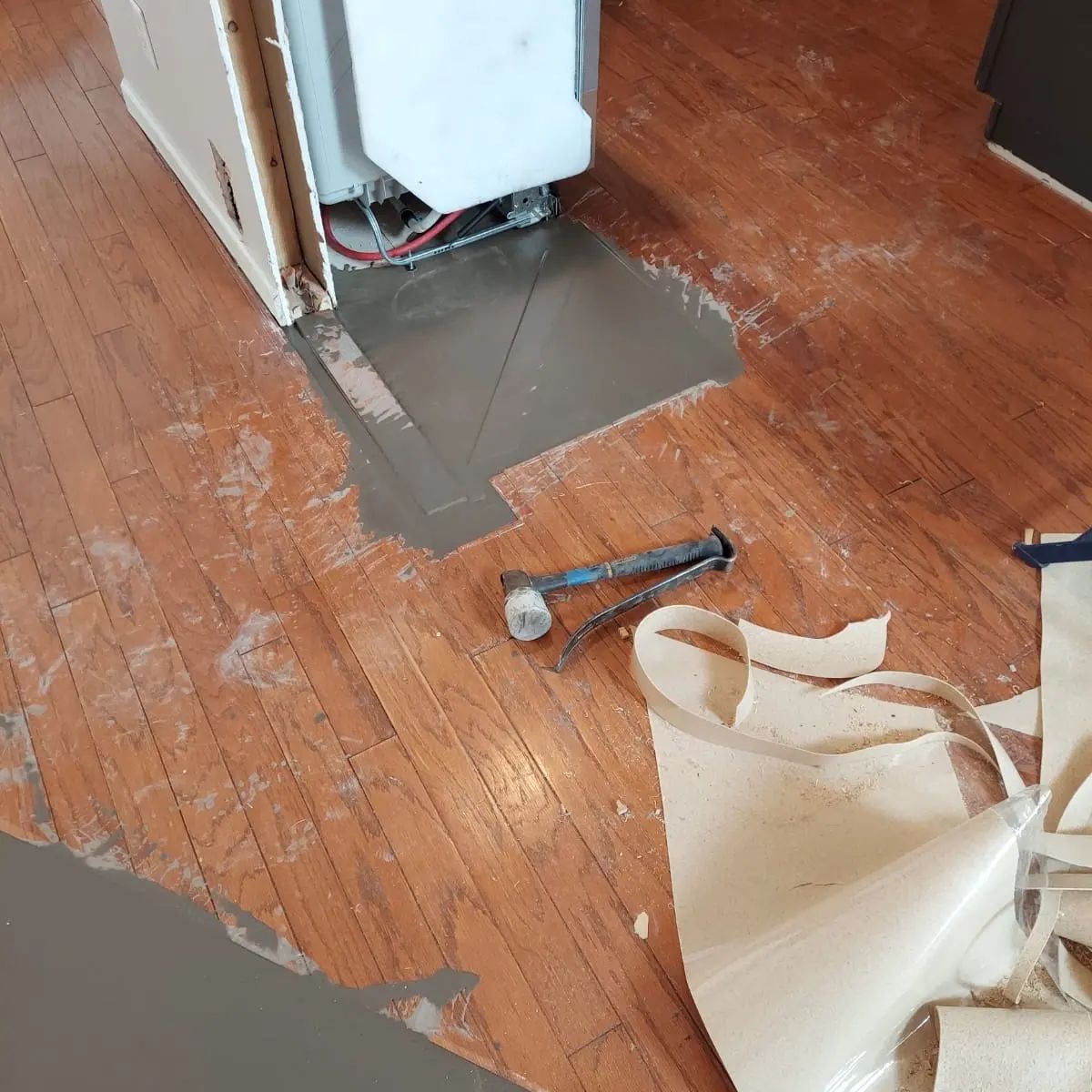 Wooden floor with a freshly poured patch of gray leveling compound near an appliance, surrounded by scattered linoleum flooring and tools.