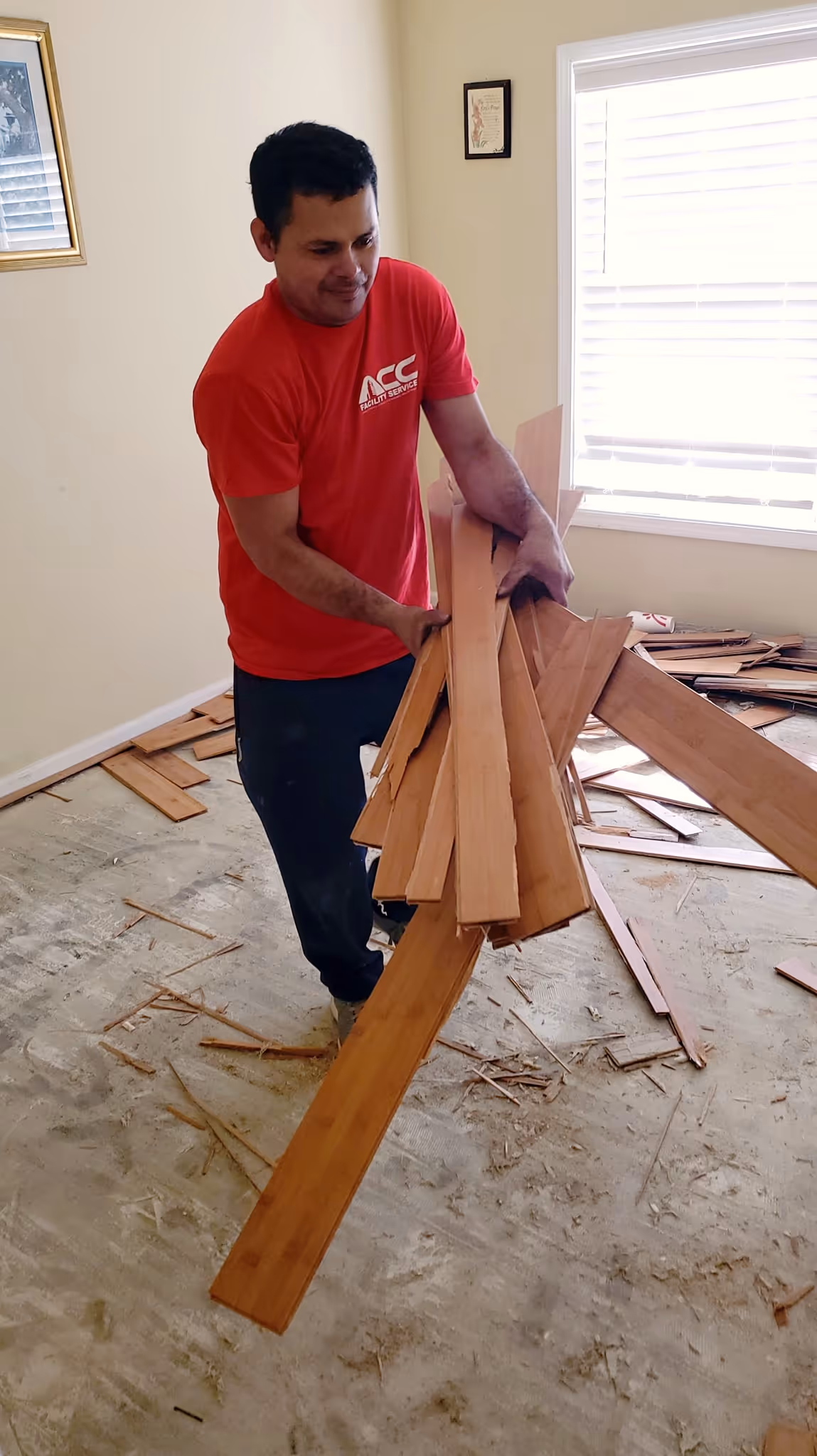 Man in red shirt holding removed hardwood floor planks in a room under renovation.