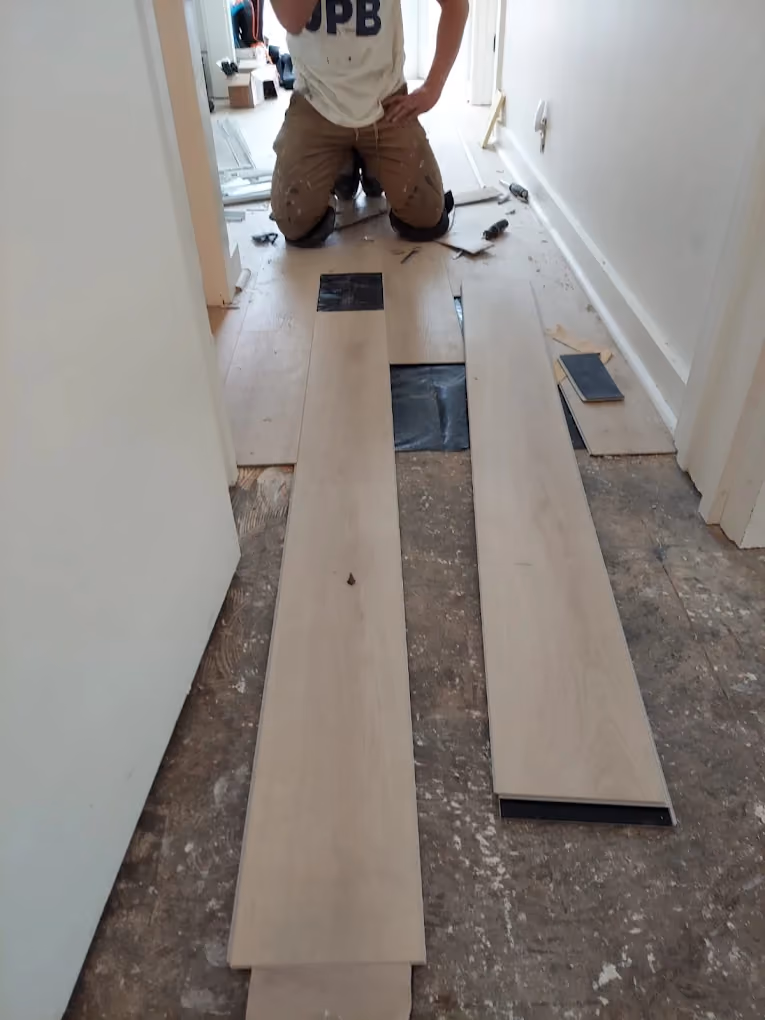 Worker kneeling installing light wood plank flooring over subfloor in a narrow hallway.