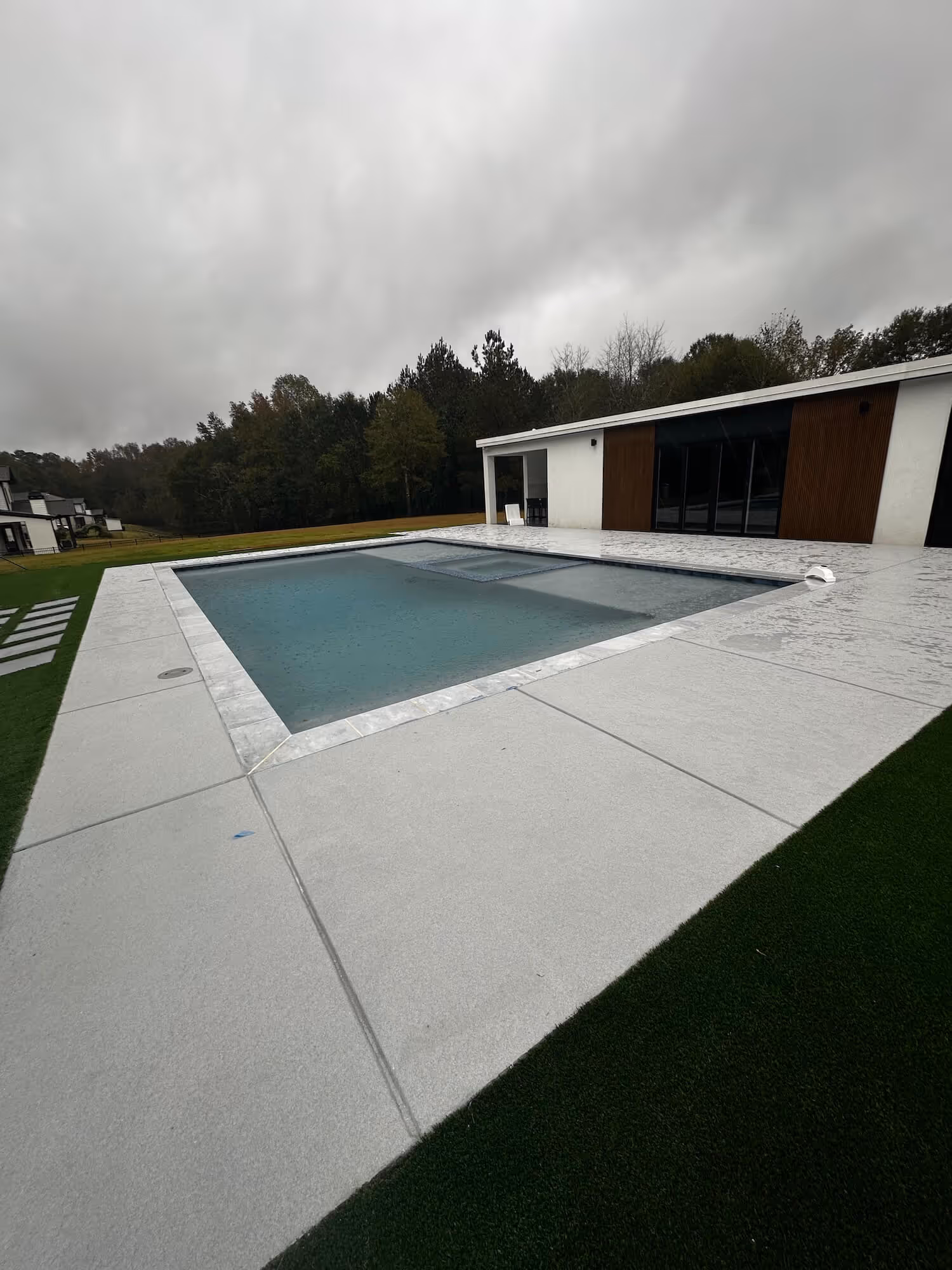Modern rectangular swimming pool with clear blue water surrounded by light gray concrete deck and artificial grass under a cloudy sky.