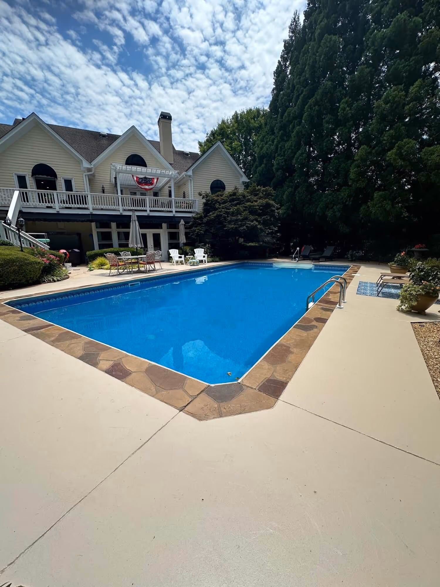 Rectangular outdoor swimming pool with blue water, surrounded by beige concrete deck and stone border, adjacent to a large house and landscaped garden.