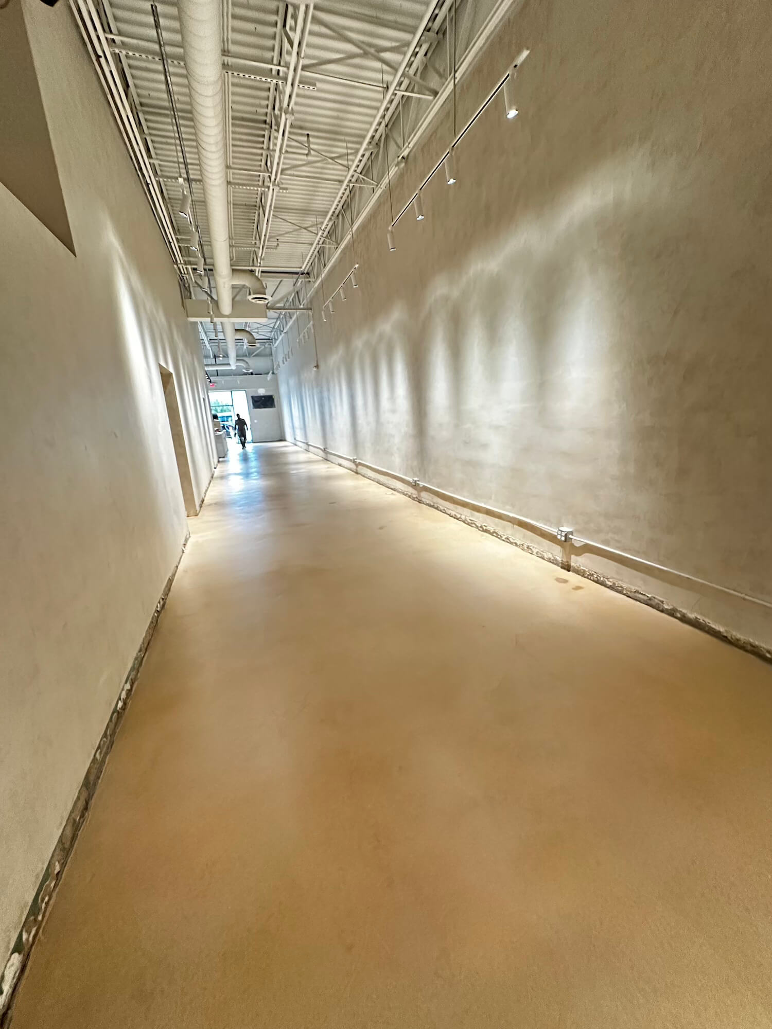 Long empty corridor with microcement beige floor, high ceiling with exposed pipes, and wall-mounted lights casting shadows.