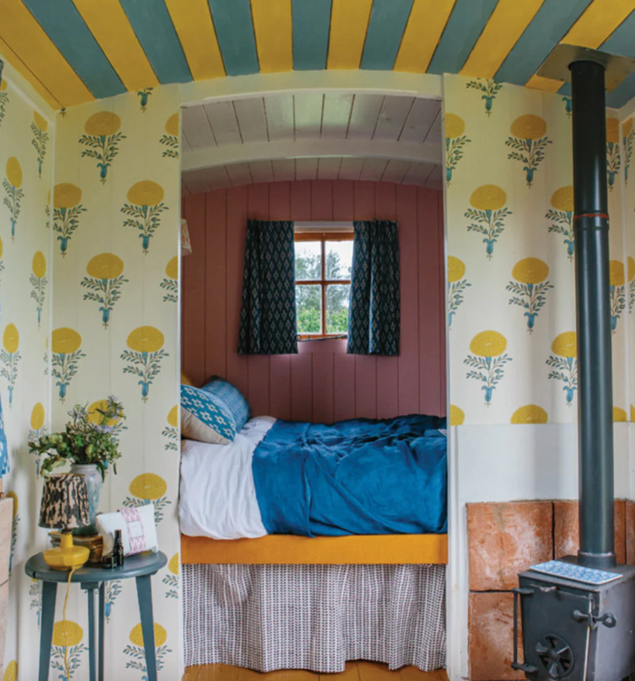 Cozy bedroom with floral wallpaper, blue-striped ceiling, wood stove, and window curtains.