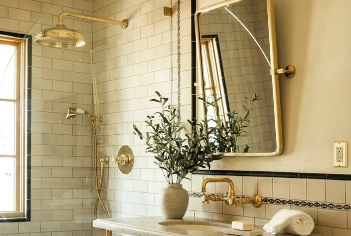 Bathroom with brass fixtures, wall mirror, potted plant, and white tiled walls.