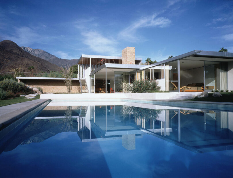 Modern house with large glass windows and a stone chimney, beautifully reflected in a clear outdoor swimming pool—this Friday Five pick stands against majestic mountains and a blue sky.