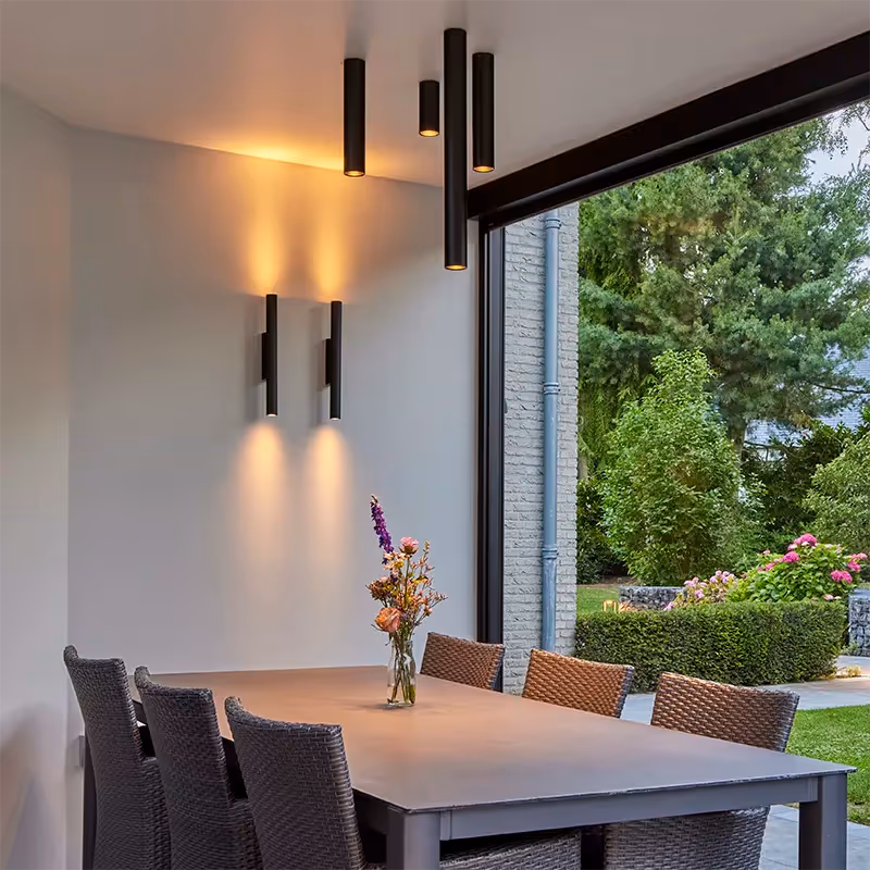 Sfeerverlichting voor buitentafel, bestaande uit slanke ‘up & down’ wandverlichting en in lengte variërende, buisvormige plafondarmaturen.