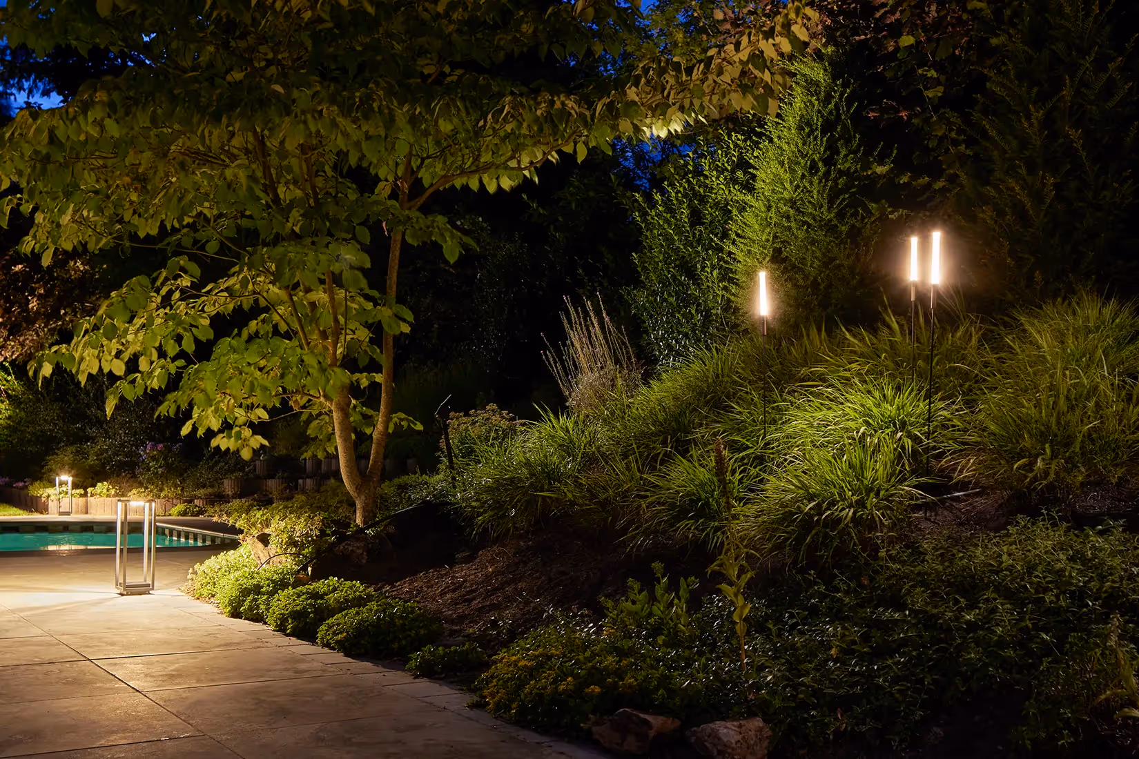 Tuin met kubusgevormde vloerarmaturen en minimalistische grondarmaturen op lange standaard met elegant lichteffect.