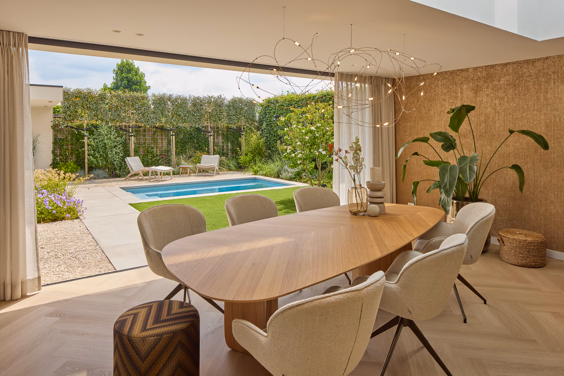 Hangarmaturen met organische vorm boven een houten eettafel met uitzicht op de tuin met zwembad.