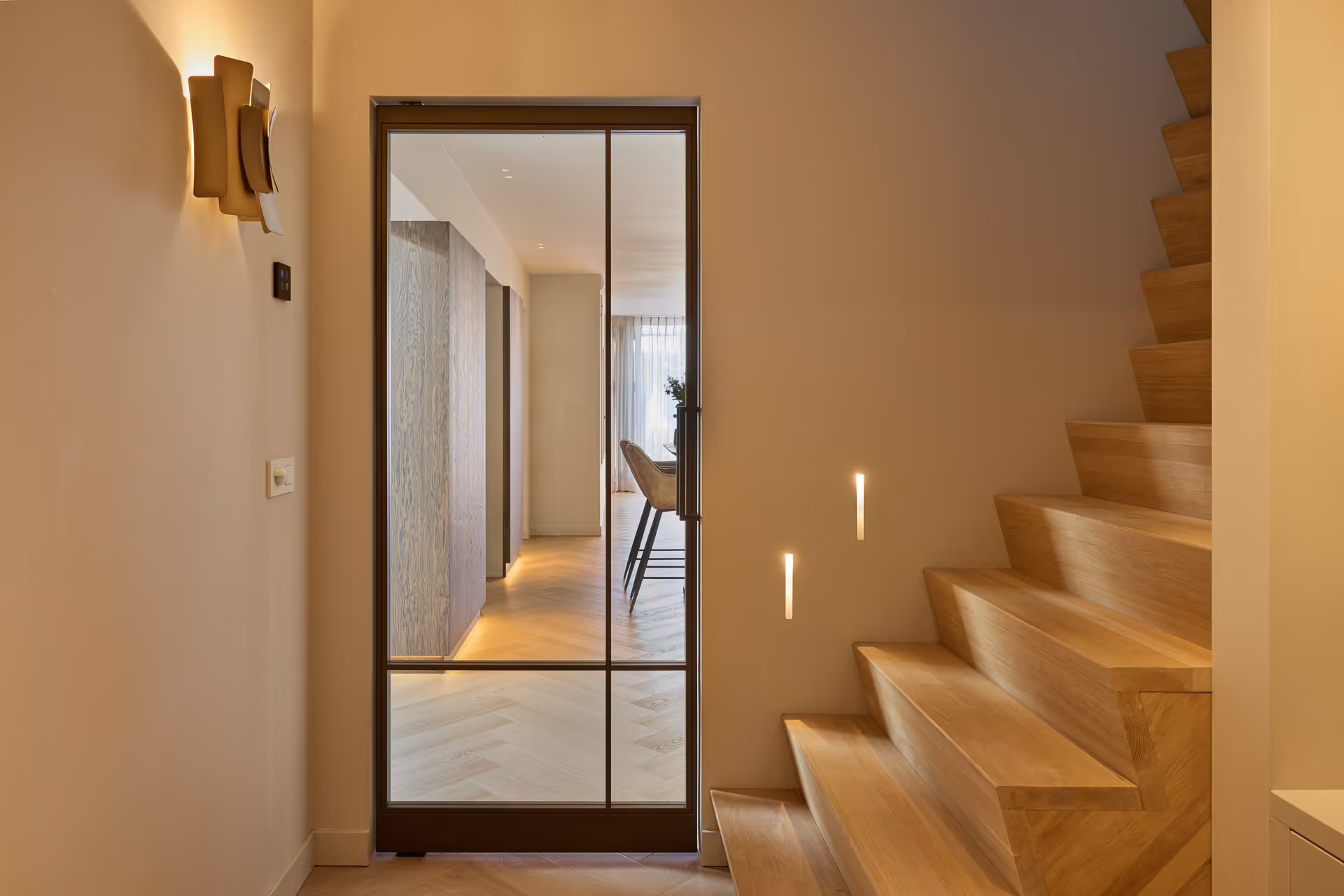 Warme hal met messing wandarmatuur en subtiele trapverlichting die de houten treden accentueert richting woonkamer.