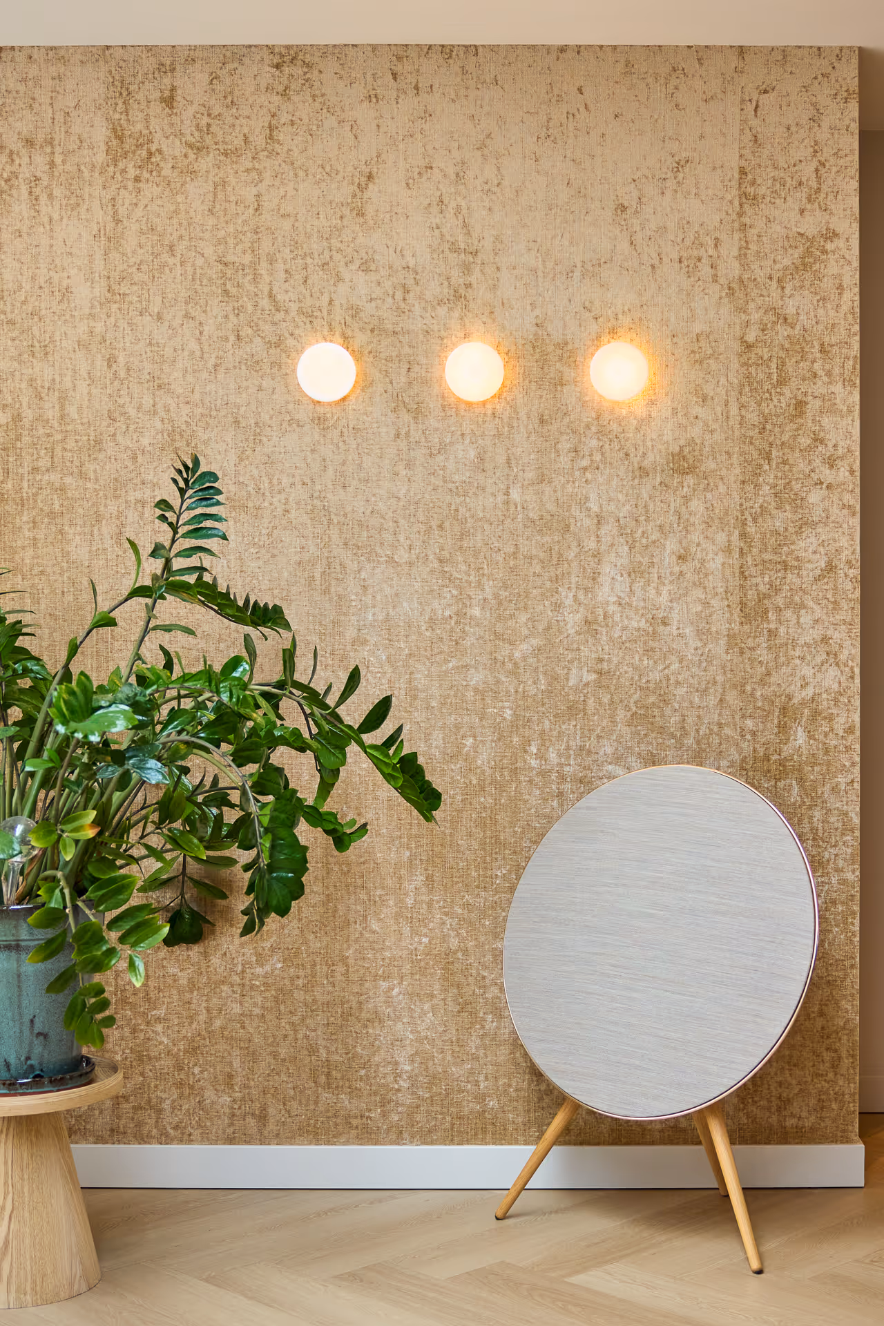 Drie cirkelvormige wandlampen verspreiden zacht diffuus licht boven goudgetinte wand met plant.