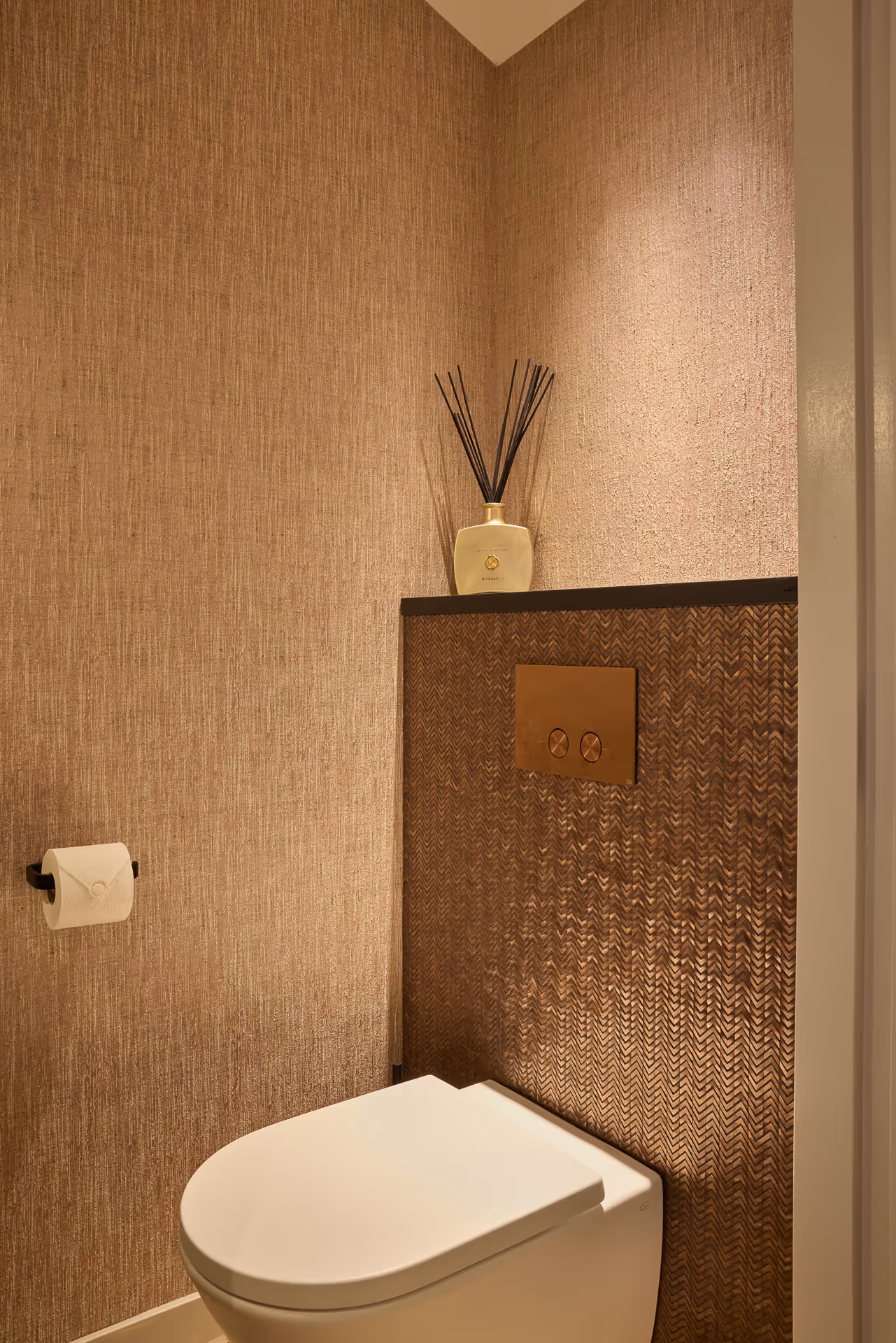Warm verlichte toiletruimte met bronskleurige wandtextuur en subtiele lichtinval boven toilet.