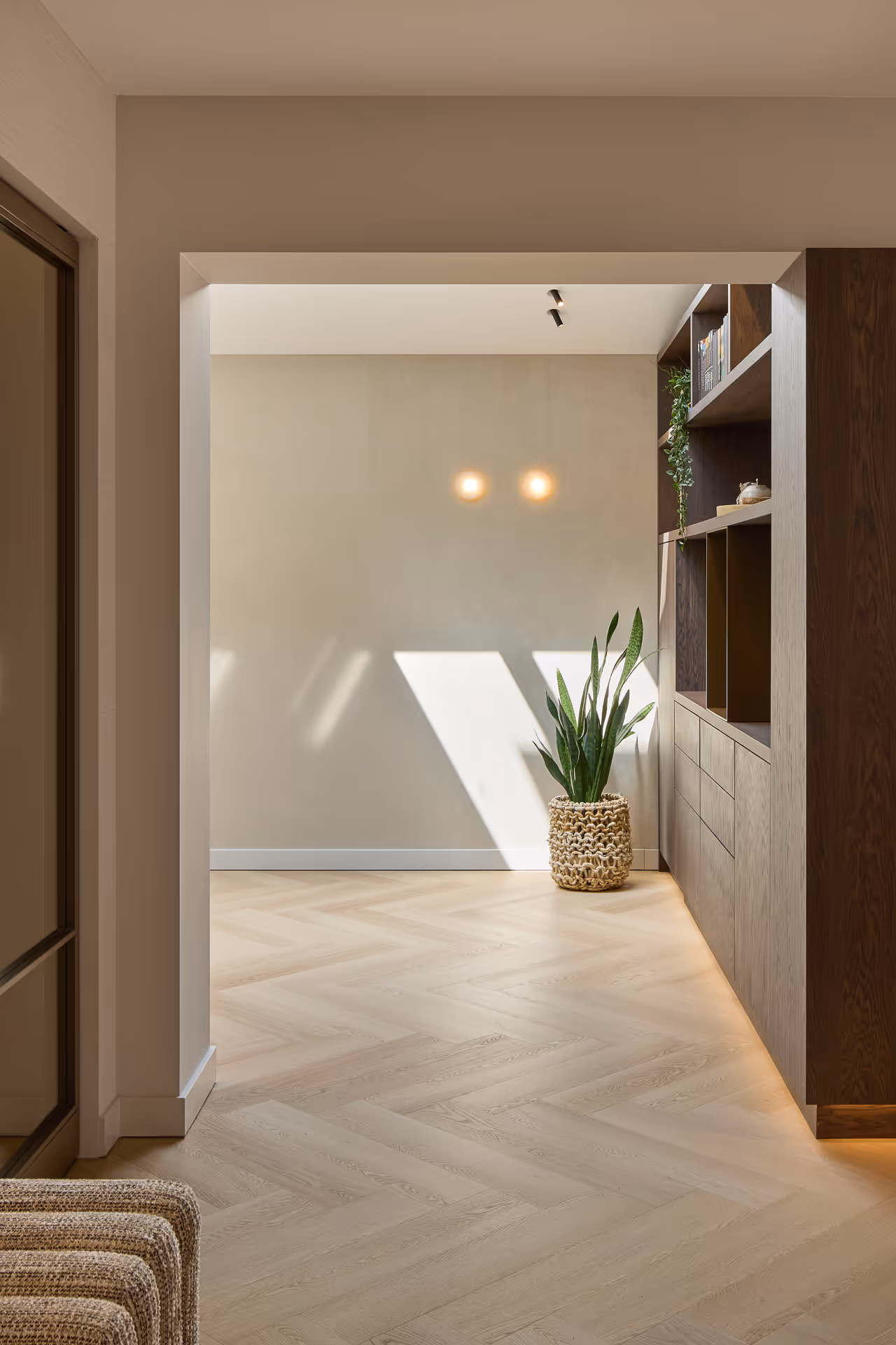 Doorkijk naar werkkamer met houten wandkast met open vakken, subtiel verlicht door ronde wandarmaturen en richtspots.