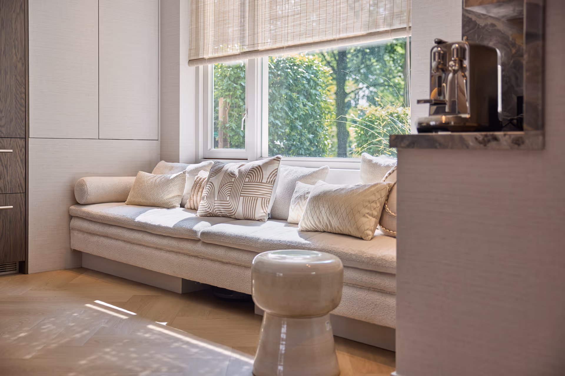 Lichte zitbank bij raam met neutrale stoffen en kussens, omhuld door natuurlijk daglicht.