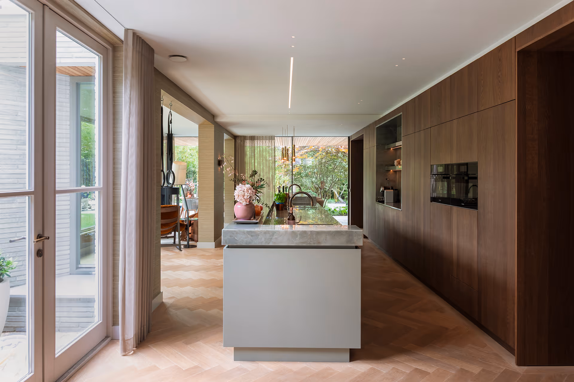 Overzicht van moderne keuken met lichtlijn in plafond, centraal eiland en houten wandafwerking in natuurlijke tinten.