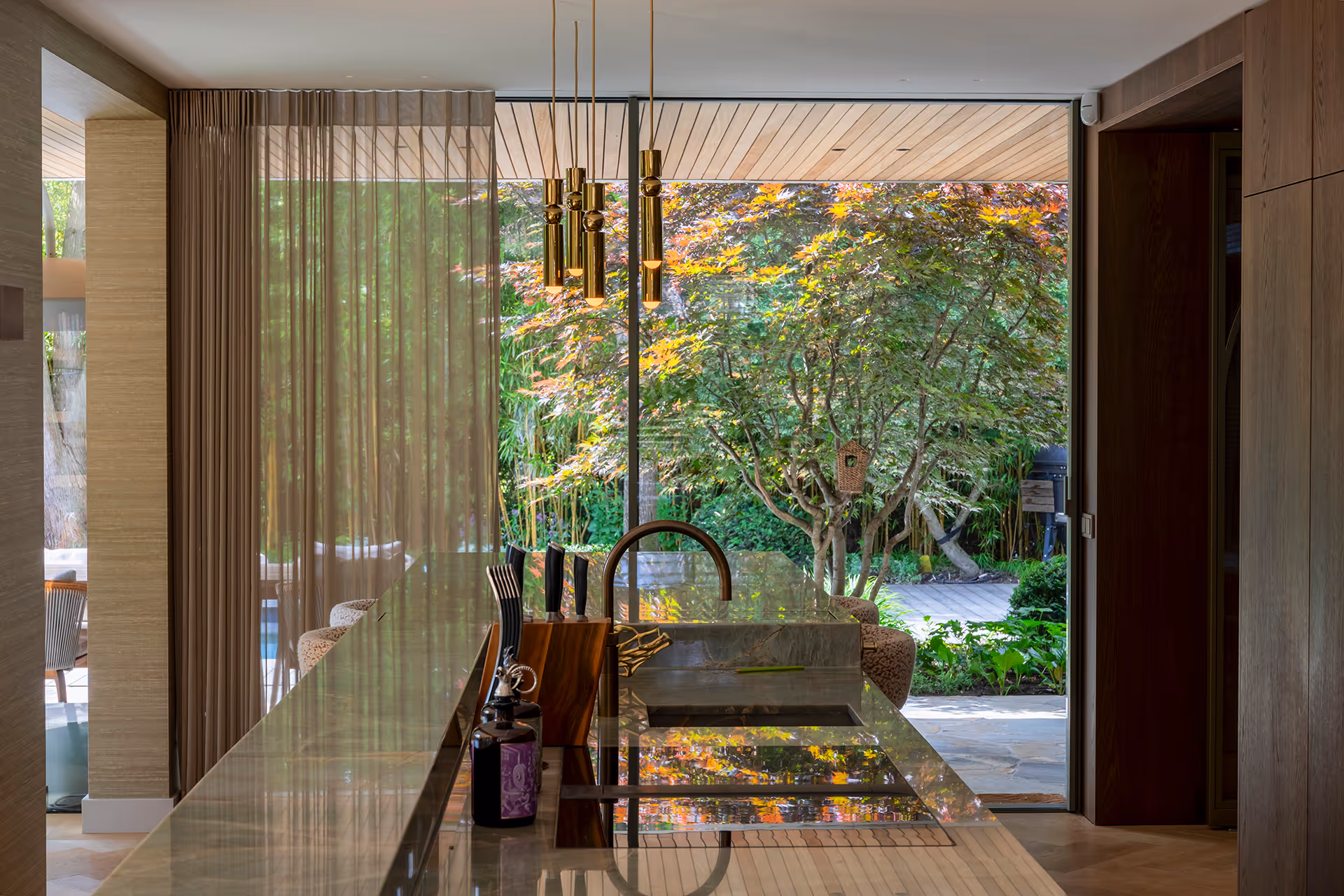 Messing hanglampen boven keukeneiland, met uitzicht op tuin door hoge glazen schuifpui die overvloedig daglicht binnenlaat.