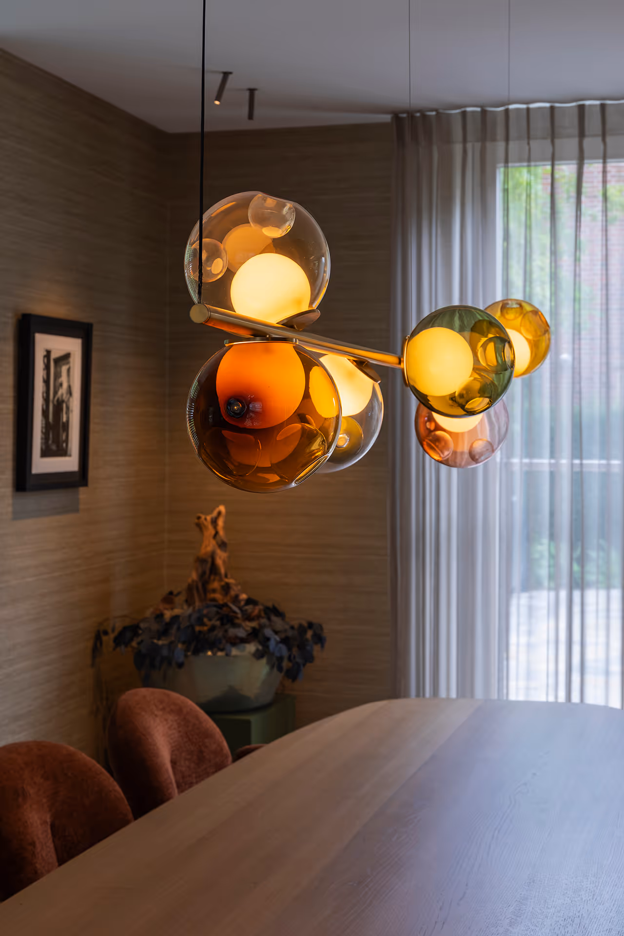 Clusterlamp van gekleurde glasbollen boven houten eettafel met zacht gefilterd daglicht door transparante gordijnen.