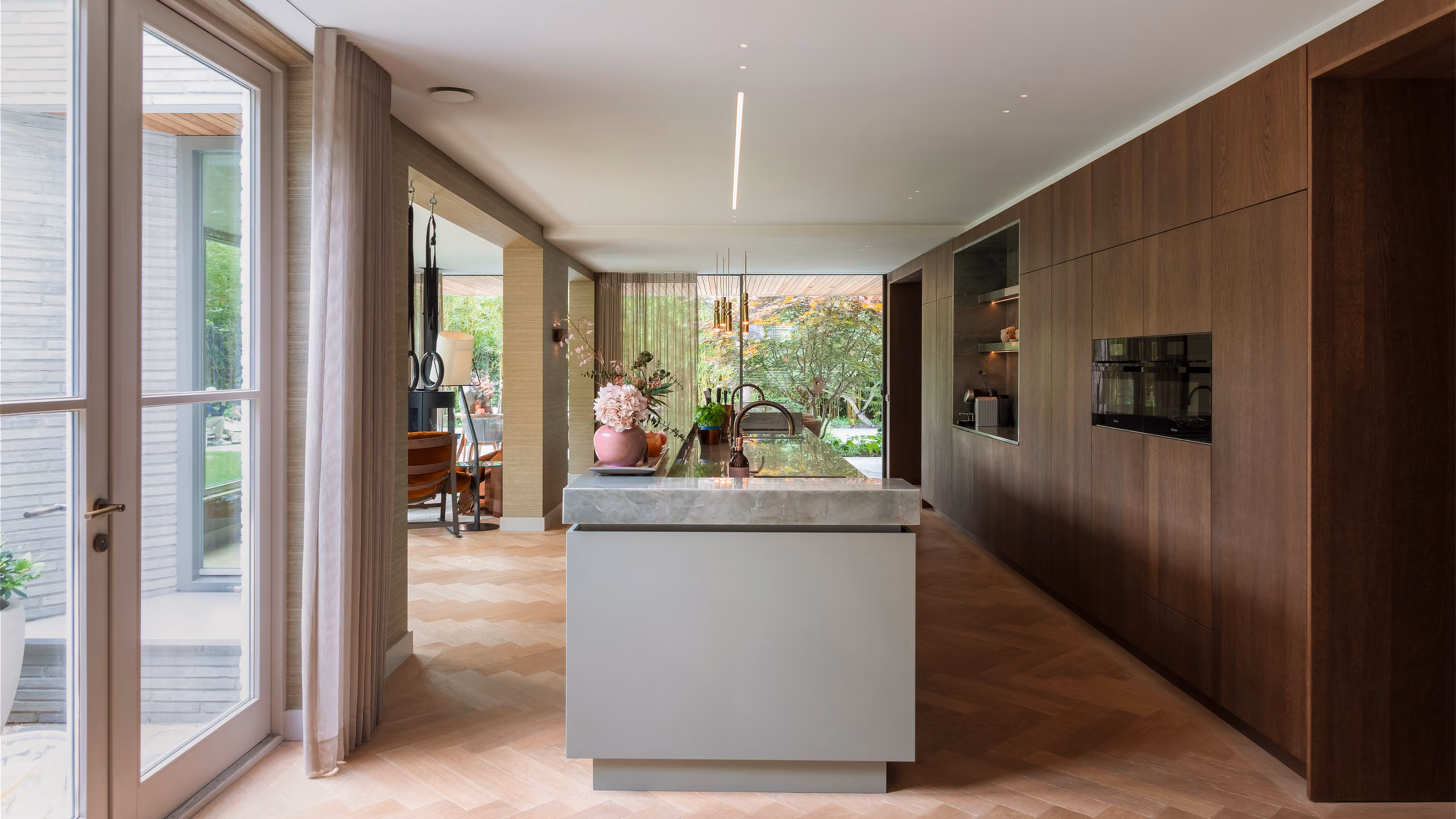 Overzicht van moderne keuken met lichtlijn in plafond, centraal eiland en houten wandafwerking in natuurlijke tinten.