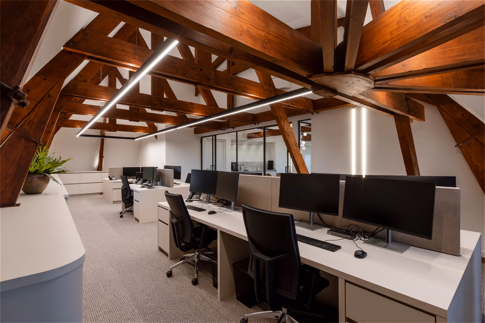Werkplekken met dubbele beeldschermen onder een lineaire hanglamp tegen de houten kapconstructie.