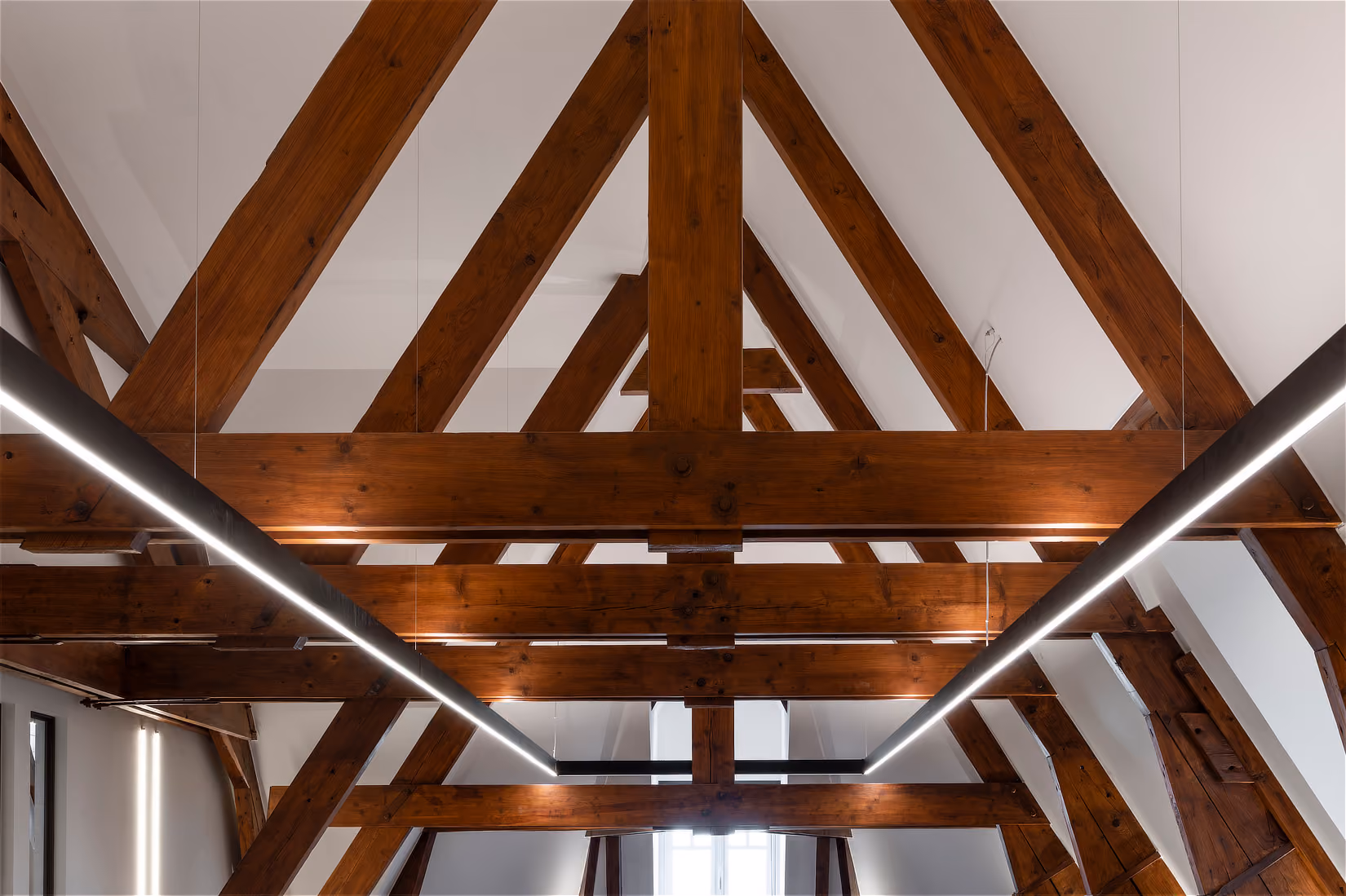 Houten kapconstructie met gehoekte spanten, geaccentueerd door kruisende lineaire verlichting.