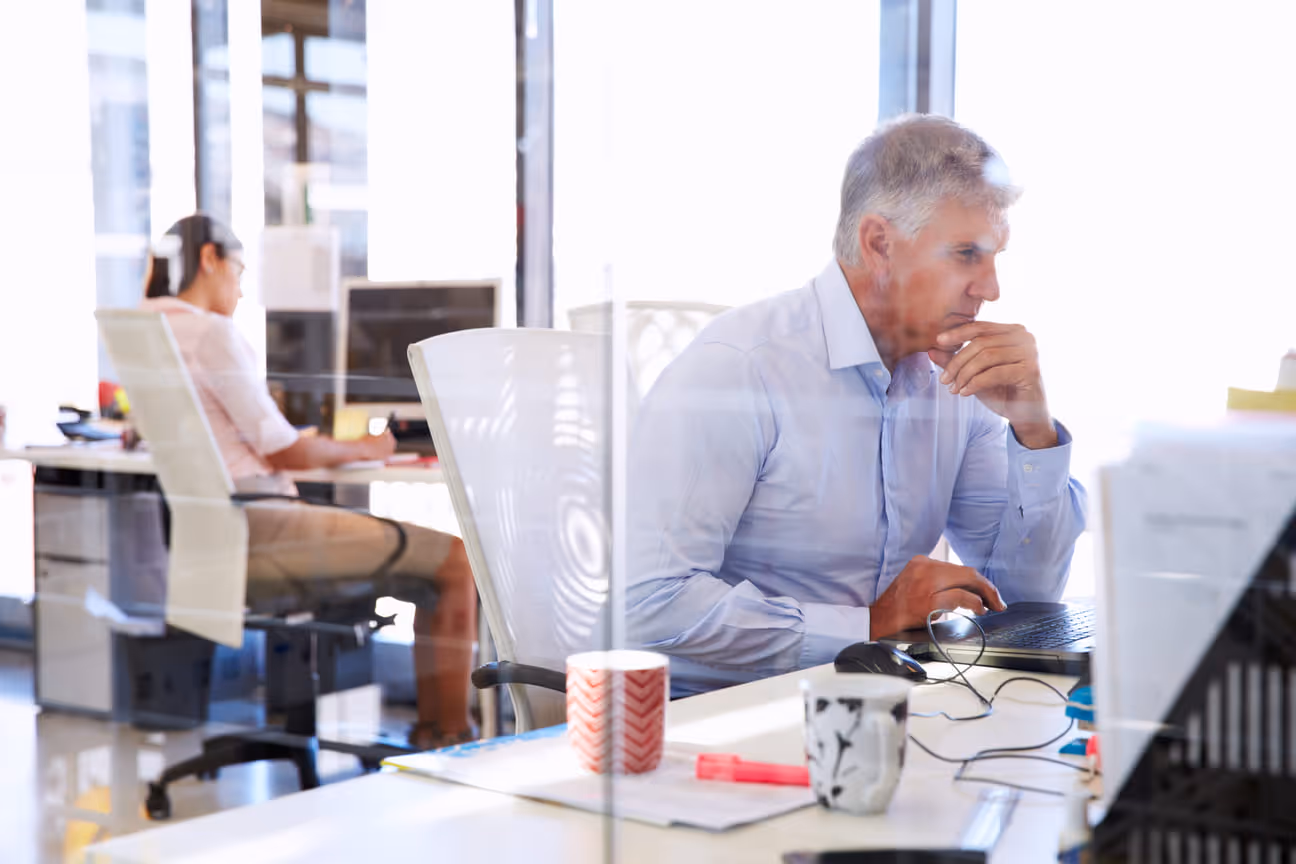 Office scene viewed through glass, showing a professional working intently at a laptop in the foreground while another colleague works at a desk in the background.