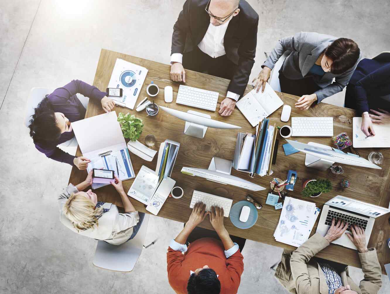 Overhead view of a business team gathered around a conference table with computers, printed charts, notebooks, and coffee, representing collaborative product planning and architecture decision-making.