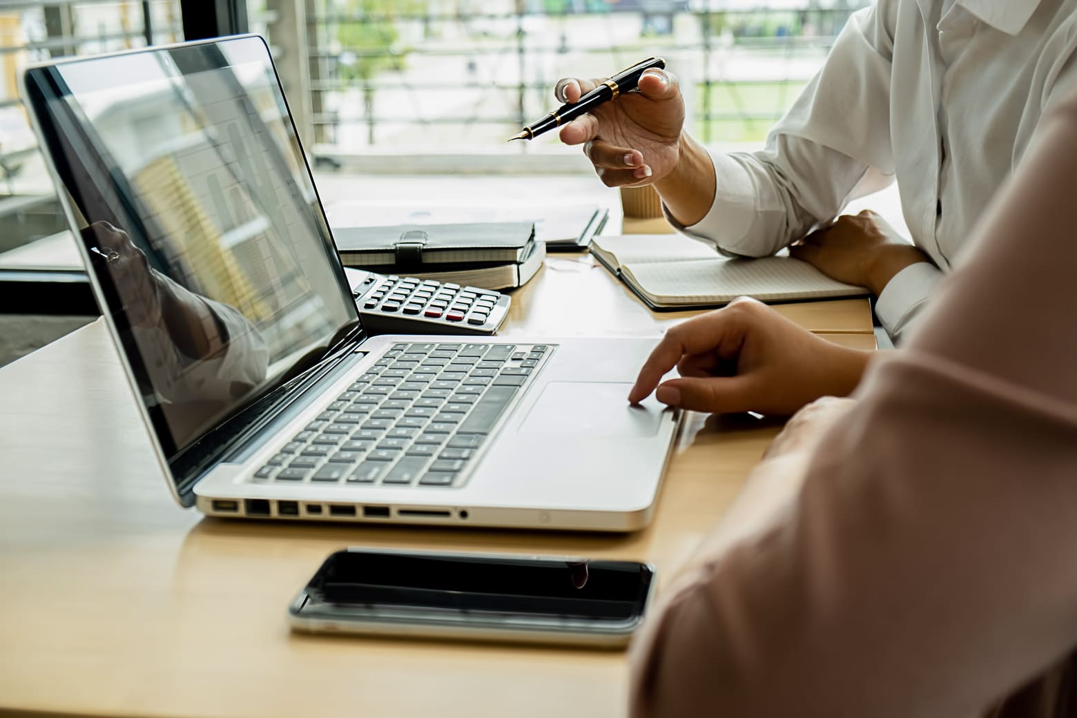 A professional working at a laptop on a wooden desk, gesturing with a pen while reviewing data, with a calculator, notebooks, and a smartphone nearby