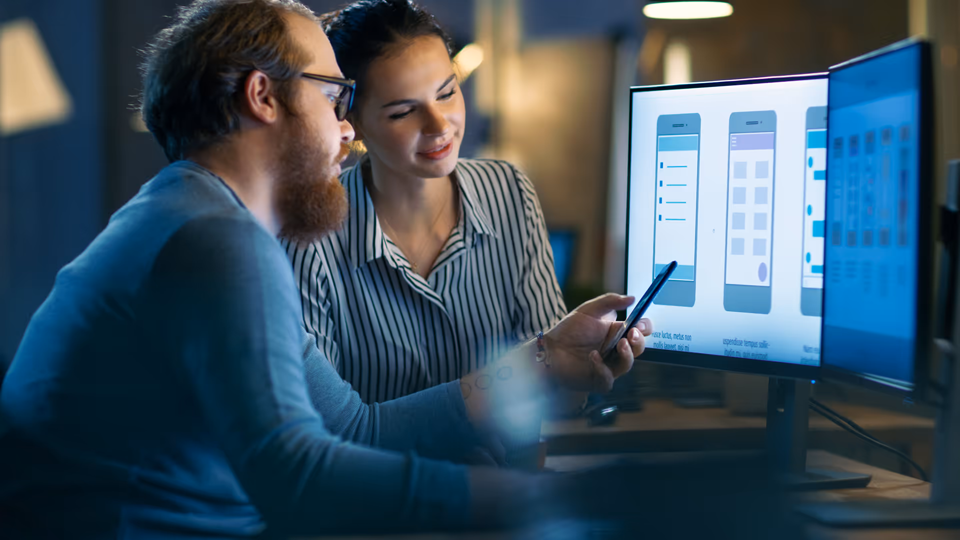 Two people reviewing mobile app wireframes on a monitor, one holding a smartphone.