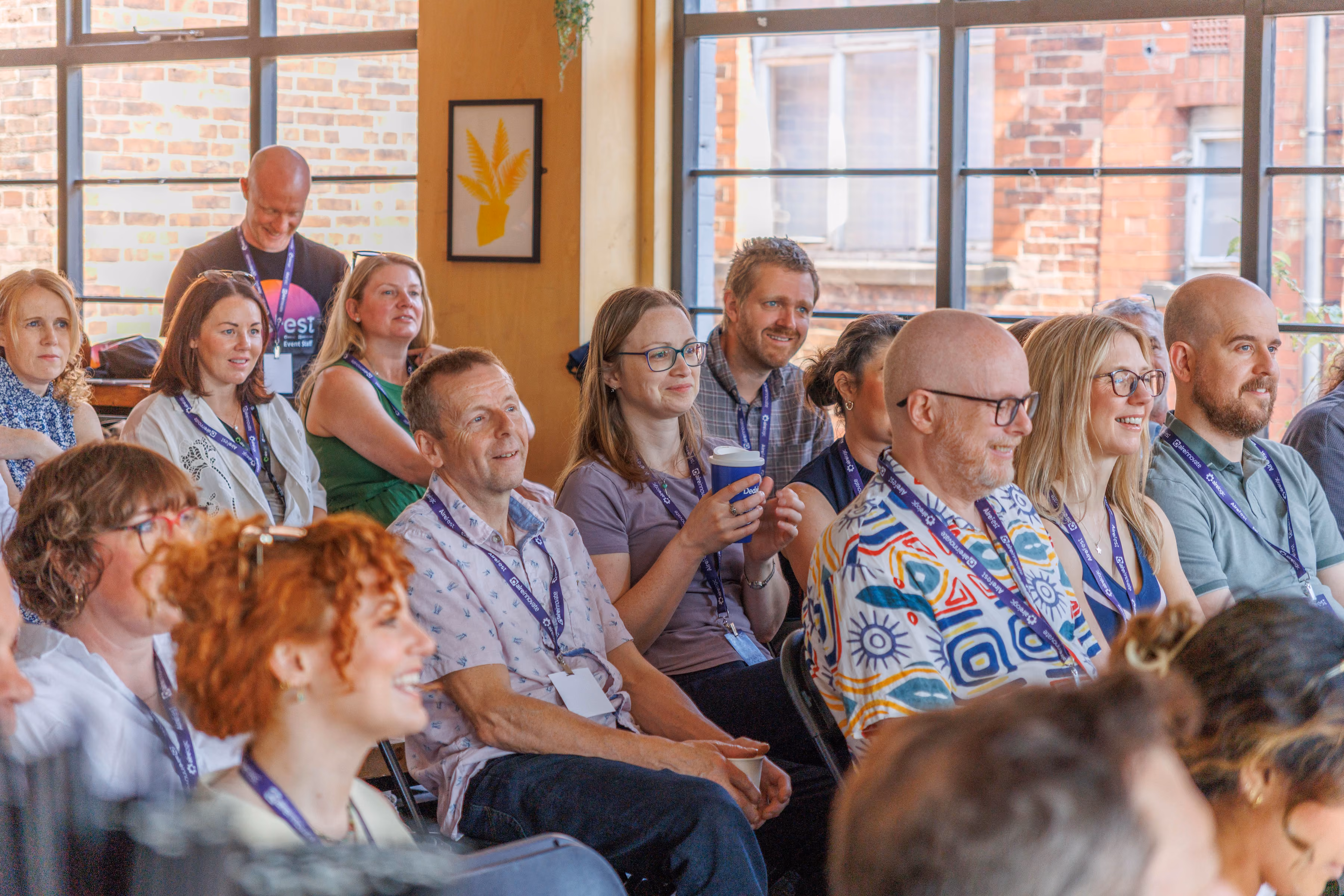 Group of adults seated indoors attentively watching an event, some smiling and holding cups.