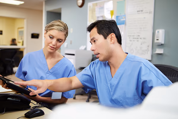 Two clinicians in blue scrubs looking at a computer screen and tablet in a medical office.